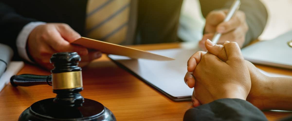 A close-up of a legal consultation scene, where a person in a suit and striped tie is holding a document while another individual sits across the desk with clasped hands. A judge's gavel is in the foreground, placed on a wooden table along with legal paperwork and a pen. The warm lighting suggests a formal or serious discussion, possibly about a legal case or settlement.